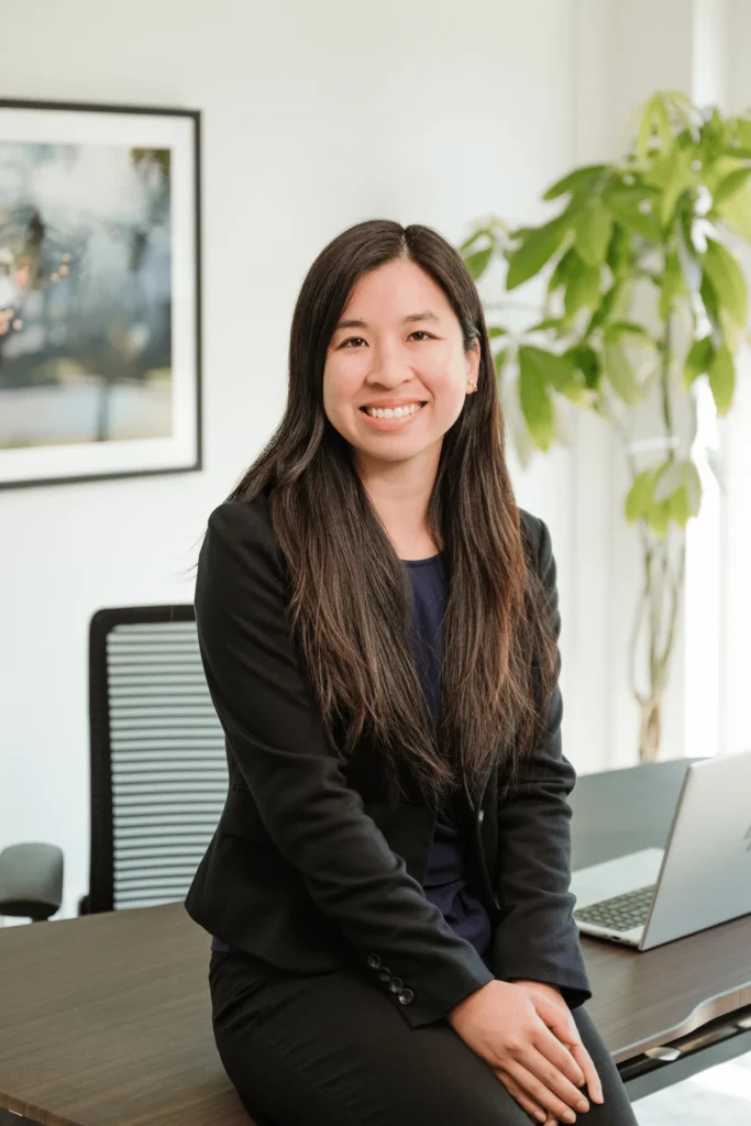 Gianna Nguyen, a CPA with long dark hair, sits confidently on a desk in her office, dressed in a black blazer and dark pants. With a laptop nearby and artwork gracing the wall behind her, she smiles warmly at the camera, exuding professionalism and charm amidst the greenery.