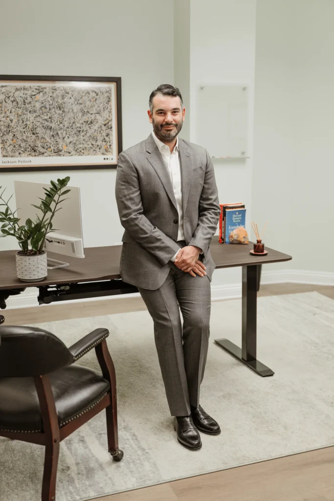 A man in a gray suit sits on the edge of a desk in an Nguyen & Company office. The desk features a computer, plant, and books. A framed map hangs on the light-colored wall behind him, subtly reflecting the CPA ethos. A black leather chair is in the foreground.