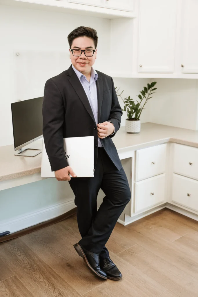 A person in a dark suit leans casually against a counter in the Nguyen & Company office, holding a closed laptop. A monitor and potted plant are visible in the background. The atmosphere is professional and modern, perfectly suited for a CPA at work.