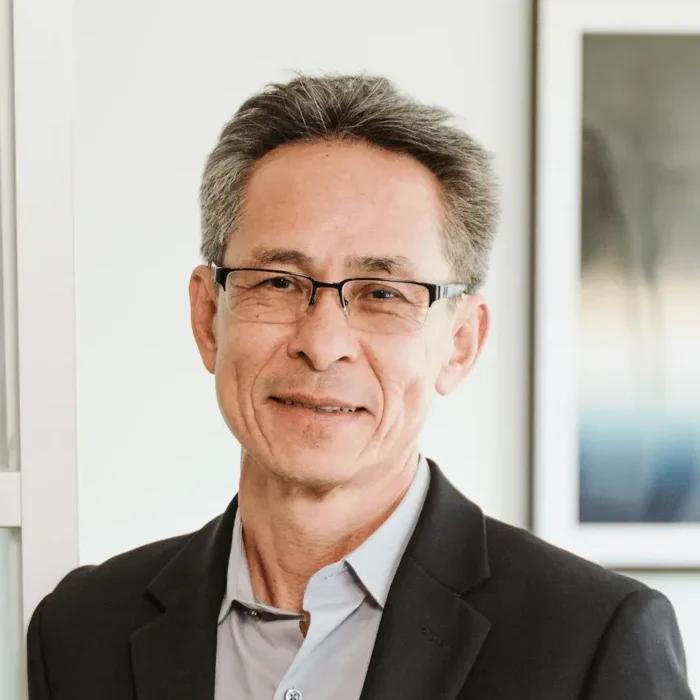 A man, likely associated with Nguyen & Company, wearing glasses and a dark blazer stands indoors, smiling slightly. He has short, graying hair and is positioned in front of a blurred picture frame on the wall, embodying the professional aura of a seasoned CPA.