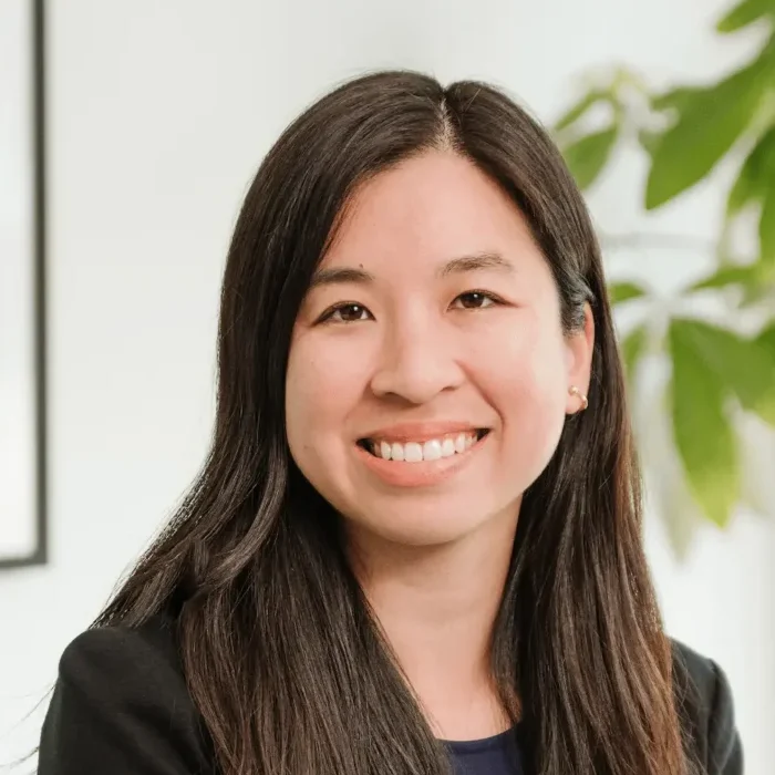 Gianna, Director of Accounting & Operational Advisory, smiles while facing the camera, exuding the confidence of a CPA. She is wearing a dark blazer and a blue top. The background is softly blurred, with some green leaves visible on the right side, hinting at her association with Nguyen & Company.