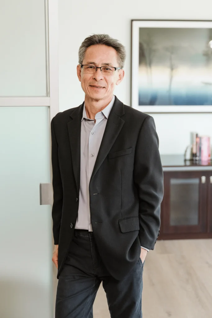 A man in a black blazer and glasses stands casually indoors, leaning against a partition at Nguyen & Company. A framed abstract artwork and some books are visible in the background, hinting at the creative yet professional environment of this CPA firm.