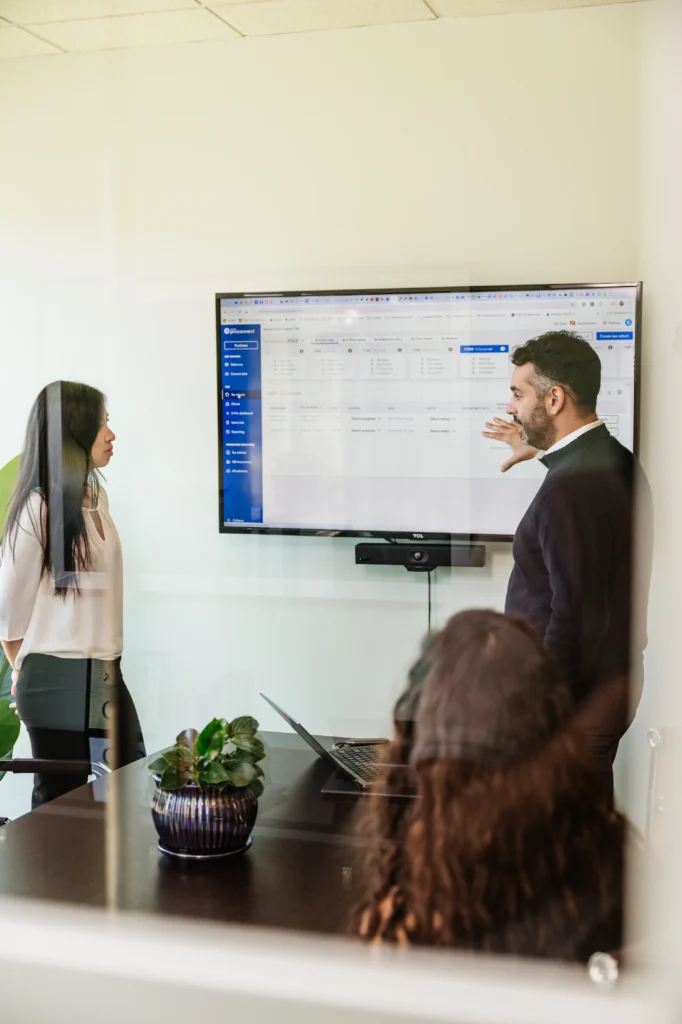 Carlos stands next to a large screen displaying a spreadsheet, gesturing towards it, while a woman listens intently. In the modern office of Nguyen & Company, with its glass wall and plant on the table, another person is seated. The atmosphere suggests a detailed CPA consultation.