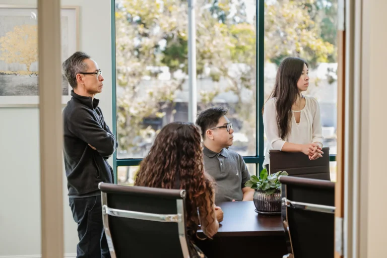 Four people in a meeting room, two standing and two seated around a table, are attentively focused on something off-camera. The professional setting of Nguyen & Company is accentuated by a plant and large windows revealing trees outside.