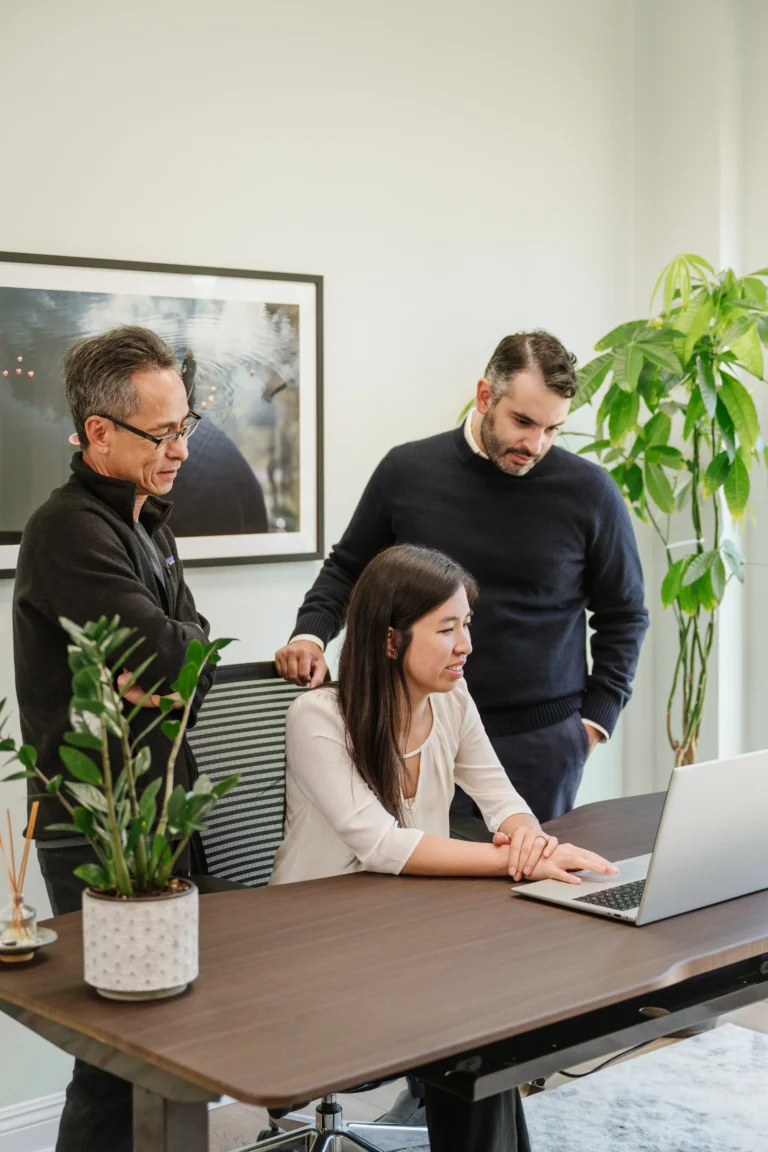 Three people are gathered around a laptop in the Nguyen & Company office. A woman, possibly the CPA, is seated and working at the computer while two men stand beside her, attentively looking at the screen. The room features a large green plant and framed artwork on the wall.