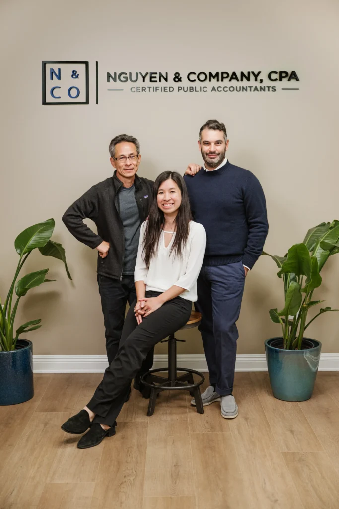 Three people are posing in an office setting. A woman sits on a stool, flanked by two men standing, all smiling warmly. Behind them, a sign proudly reads "Nguyen & Company, CPA." Two potted plants add a touch of greenery to the scene.