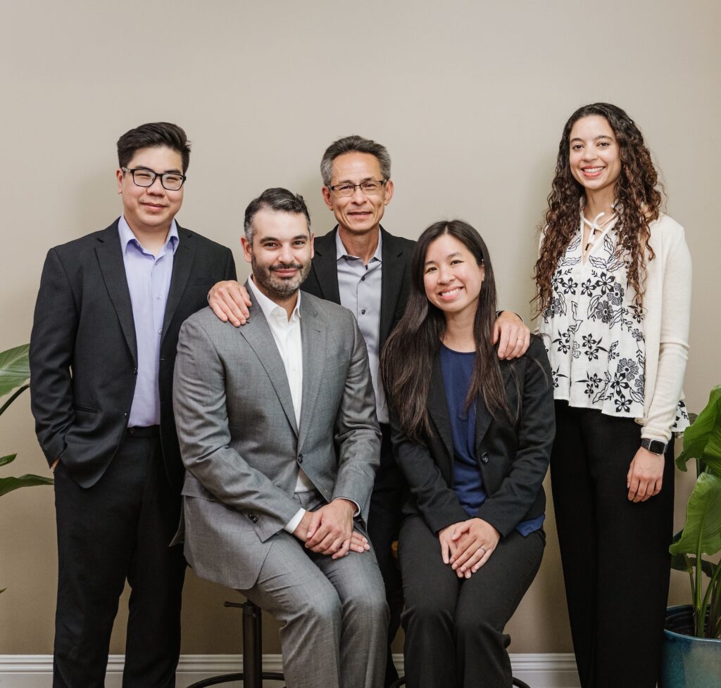 A group of five people in business attire from Nguyen & Company pose together in front of a neutral wall. Two are seated, and three stand behind them, flanked by green plants. They smile warmly at the camera, exuding a welcoming professional vibe.