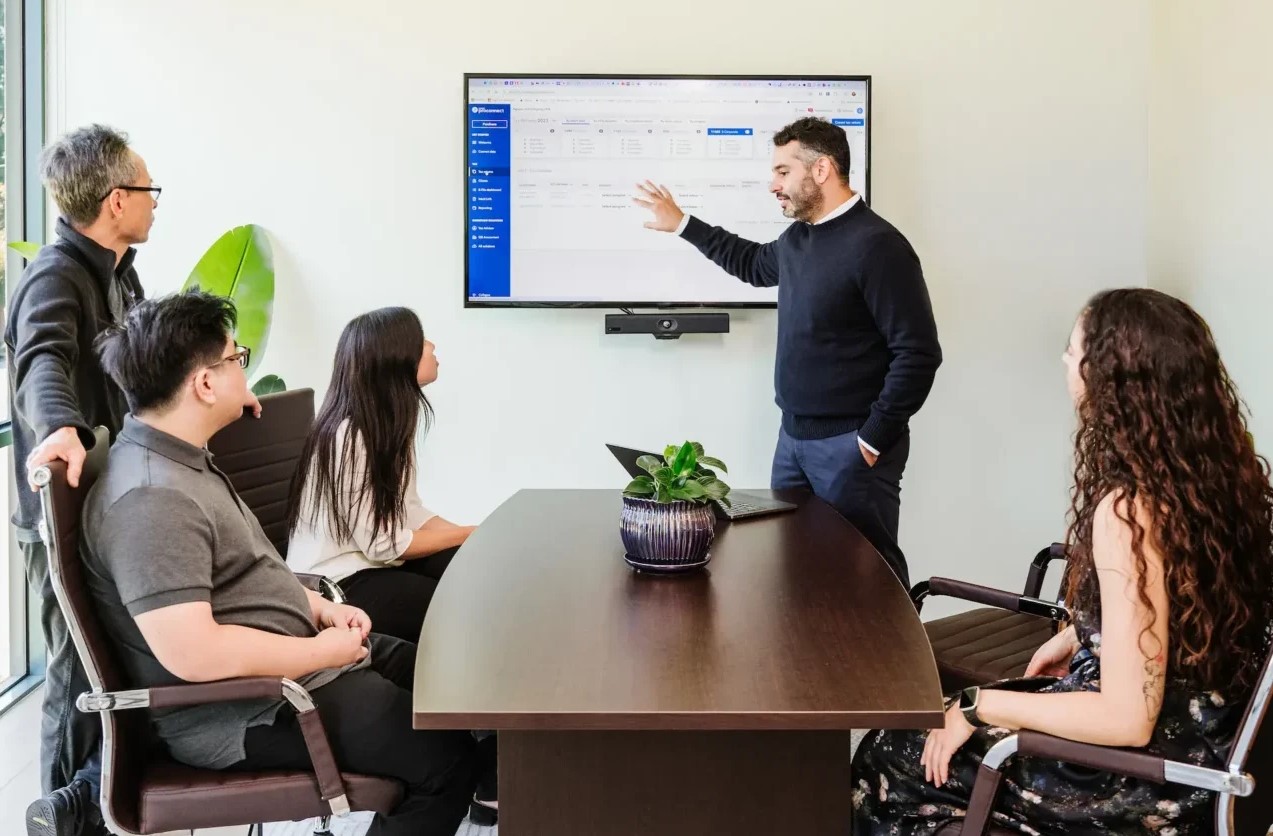 Five people are in a meeting room at Nguyen & Company. A CPA stands and points at a screen displaying a spreadsheet while four others, seated around the table, listen attentively. A laptop and plant sit on the table.